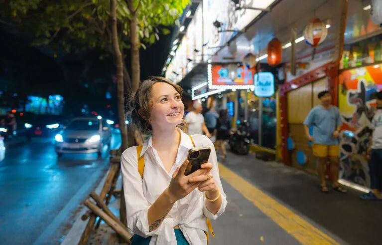 Young Caucasian woman smiles while holding with a smartphone on a street in Bangkok at night.