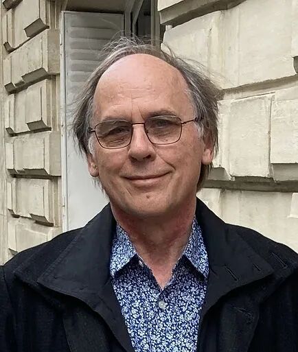 哥伦比亚大学高级讲师彼得·沃特 （Peter Woit） 戴着眼镜，穿着带图案的衬衫，在一座石头建筑前微笑。他的表情很温暖，心情很轻松。