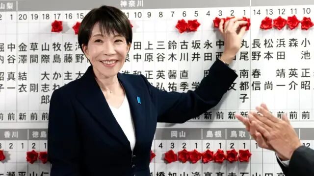 高市早苗穿着深色西装外套和白色上衣，将一枚红色胸花别在木板上。