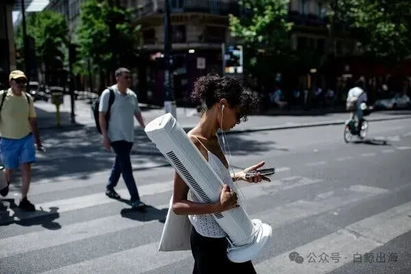 A person crosses a street, carrying a large white tower fan. They look at their smartphone; other people are in the background.