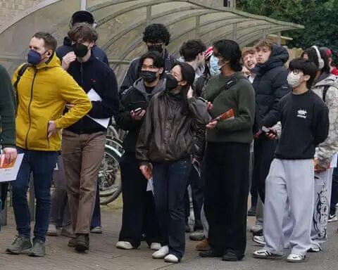 Students in face masks queue next to bikesheds.