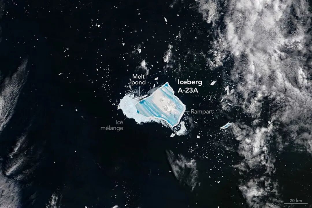 A satellite image captured on December 26, 2025, centers on iceberg A-23A adrift in dark Southern Ocean waters. The iceberg appears as a bright white, roughly rectangular slab with jagged edges and parallel blue stripes across its surface. The blue areas are meltwater ponds. A thick patch of ice fragments known as brash ice is visible along one edge of the main iceberg in an area where meltwater is leaking from it, while patches of small icebergs and clouds are also visible around it.
