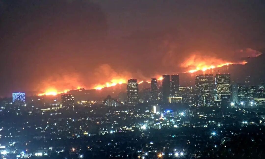 Still image from PTZ Camera on roof of high rise in downtown Los Angeles - taken of Palisades Fire at peak intensity on the evening of January 7, 2025. The city with twinkling lights and skyscrapers is in the foreground. In the background a line of wildfires burn across the middle of the image.