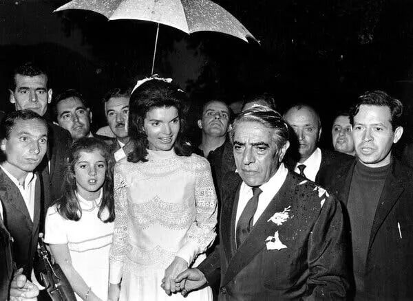 Jacqueline Kennedy wearing a white high neck dress with scalloped lace and buttons on a sleeve. She’s holding hands with Aristotle Onassis and they’re walking with a crowd behind them, someone is holding an umbrella over her head.