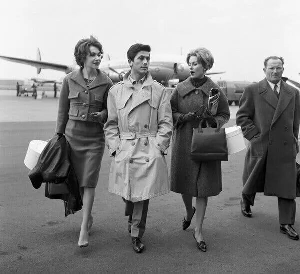 A black and white photo of Valentino wearing a light colored trench coat walking away from an airplane with two women on either side.