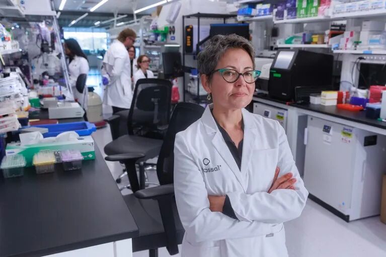 Beth Shapiro poses for a portrait in a lab with research equipment and other scientists working a