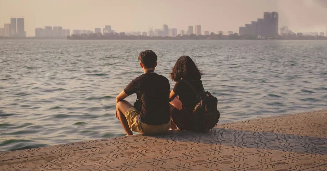 a man and woman sitting on a dock by the water with a city in the background