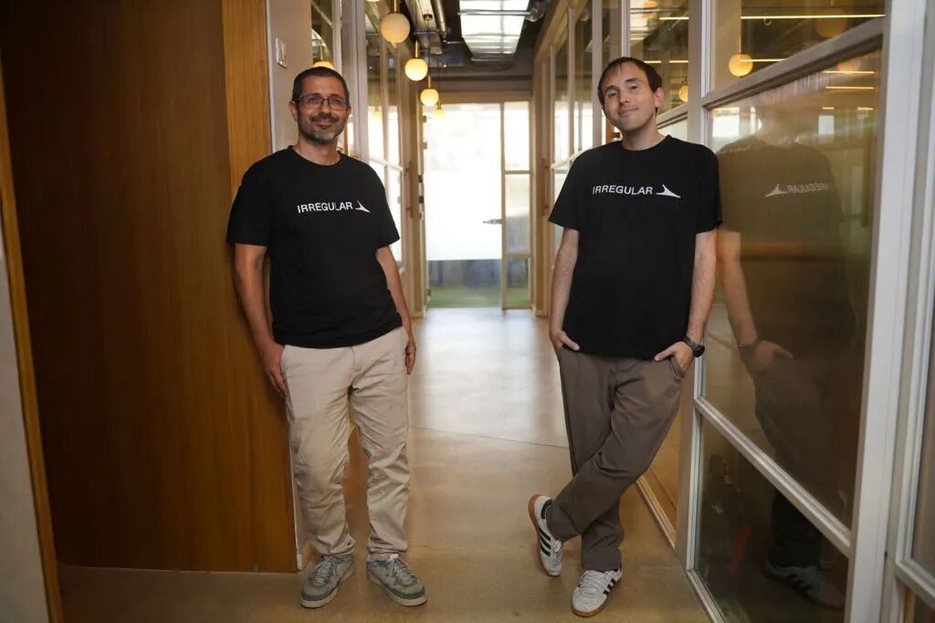 The two co-founders stand abreast in an office hallway wearing black logo t-shirts.