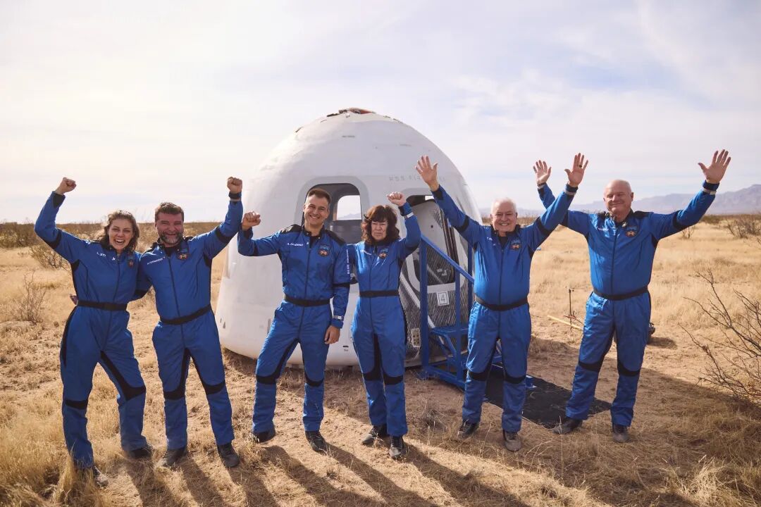 The NS-38 crew of six people in blue spacesuits cheerfully celebrating near the crew capsule, after a successful mission, set in a desert landscape.